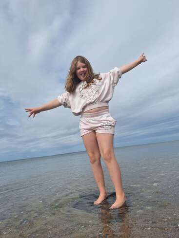 Ein junges Mädchen steht barfuß im seichten Wasser am Strand und streckt freudig die Arme aus. Sie lächelt und genießt die ruhige Atmosphäre am Meer unter einem meist bewölkten Himmel.