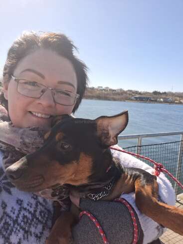 Eine lächelnde Frau mit Brille hält an einem sonnigen Tag am Ufer einen schwarz-braunen Hund im Arm. Der Hund trägt einen kuscheligen Pullover.
