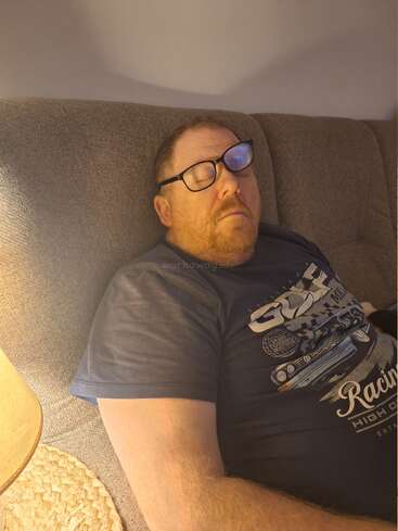 Ein Mann mit Brille schläft auf einem grauen Sofa. Er trägt ein dunkelblaues T-Shirt mit Aufdruck. Eine Lampe neben ihm taucht die gemütliche Szenerie in sanftes Licht.