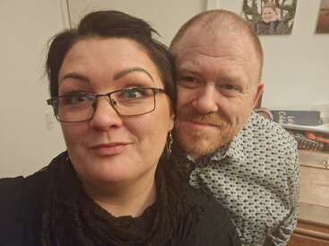 Eine Frau mit Brille und ein Mann mit Bart lächeln und posieren drinnen für ein Selfie. Im Hintergrund sind Fotos und Bücher zu sehen.
