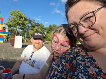 Eine Familie genießt einen sonnigen Tag im Freien. Das Kind hat ein Schmetterlings-Gesichts-Make-up, die Mutter trägt eine Brille und ein Blumenkleid, und der Vater lächelt im Hintergrund.