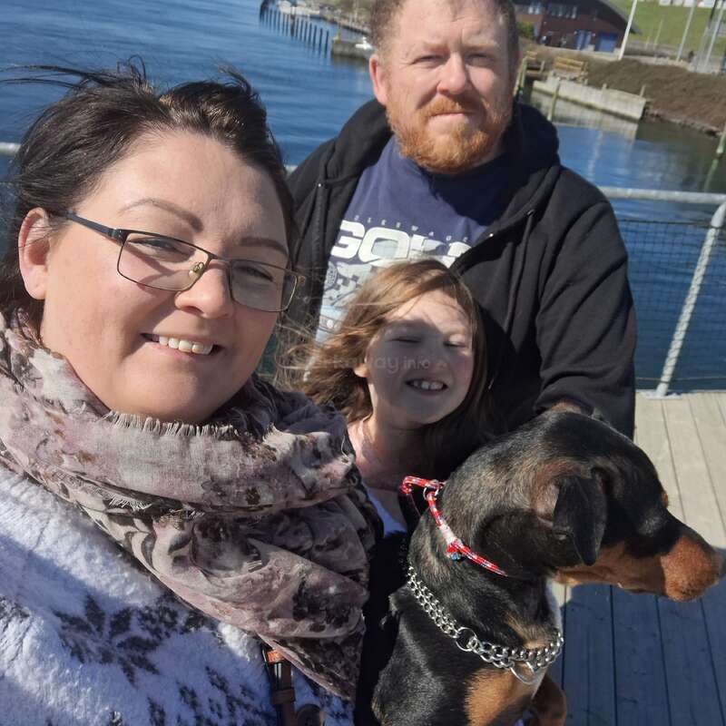Eine lächelnde Familie steht auf einem Steg am Wasser. Zu der Gruppe gehören eine Frau, ein Mann, ein Kind und ein schwarz-brauner Hund, die alle den sonnigen Tag genießen.