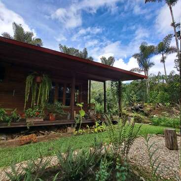 L'image représente une maison en bois avec un porche, entourée d'une végétation luxuriante et d'un ciel bleu avec des nuages blancs. La maison est située dans un environnement tropical.