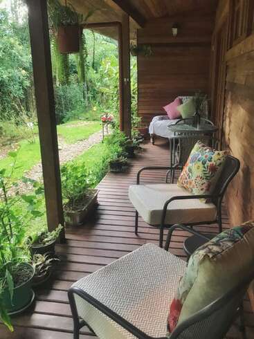 Un porche en bois confortable décoré de plantes, de chaises confortables avec des coussins colorés et d'un petit canapé, donnant sur un jardin verdoyant et un sentier tranquille.