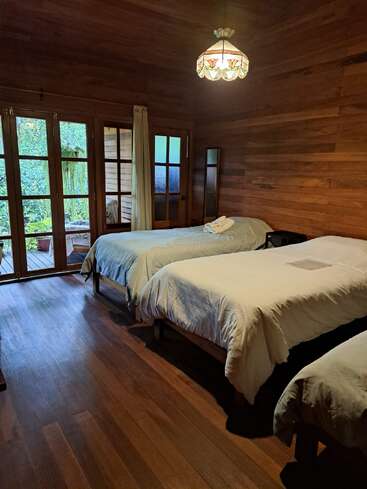 Une chambre confortable en bois avec deux lits simples, une literie blanche, un éclairage doux, des portes vitrées s'ouvrant sur la verdure, une lampe vintage et une atmosphère tranquille et rustique.