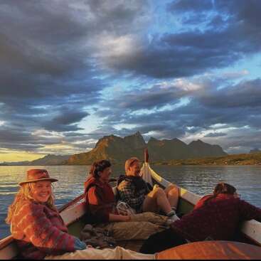 The image depicts a serene scene of four individuals in a small boat on a tranquil body of water, set against a majestic mountainous backdrop under a cloudy sky.