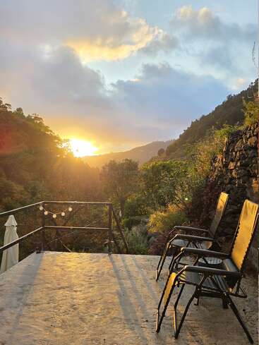 Two empty chairs face a stunning valley sunset from a concrete terrace, surrounded by lush greenery, string lights, and warm golden sunlight illuminating the scene.