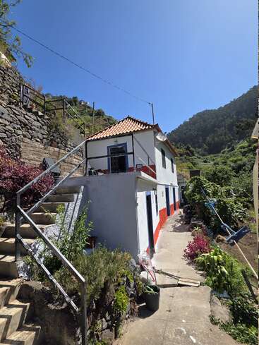 A charming white house with a red-trimmed base sits on a lush hillside. There's a stone staircase, garden plants, and mountains under a clear blue sky.