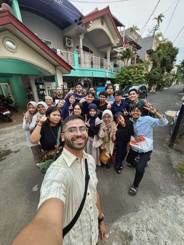 Um grupo de pessoas alegres posa para uma selfie em uma rua residencial, sorrindo e fazendo sinais de paz, com casas coloridas visíveis ao fundo.