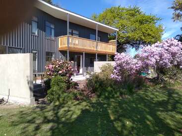 Ein modernes, zweistöckiges Haus mit einem Holzbalkon blickt auf einen üppigen Garten mit rosa Blumen und grünen Sträuchern unter einem hellen, klaren Himmel.