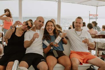 Four friends sit closely on a boat, laughing, holding drinks and a bottle. They appear to be enjoying a lively sunny day near the water.