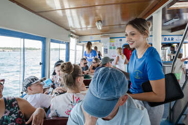 A group of people enjoy a boat ride. A smiling woman in a blue shirt interacts with passengers. Sunlight streams through windows, revealing water and scenic views outside.