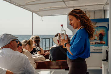 A cheerful woman in a blue shirt holds a bottle, engaging with seated passengers on a boat. The sun shines, and the sea is visible outside.