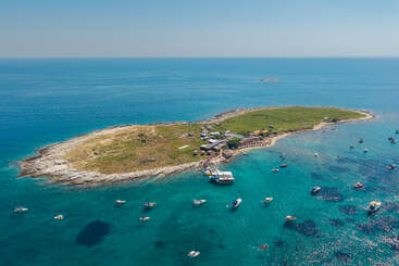 This aerial view shows a small island surrounded by clear blue water, with several boats anchored nearby. There are buildings and grassy areas on the island.