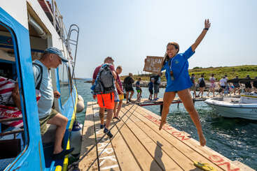 A joyful woman greets visitors disembarking a boat onto a wooden dock. Other people walk along the dock by the water on a sunny day.