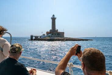 Three people on a boat photograph a stone lighthouse on a small rocky island, surrounded by sparkling blue sea under a clear, sunny sky. Peaceful scene.