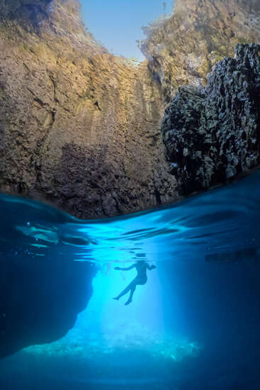 A person swims underwater in a beautiful cave, illuminated by bright blue light from above, with rocky walls surrounding the serene, crystal-clear water environment.