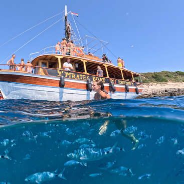 A group of people enjoy a sunny day on a wooden pirate excursion boat. A woman swims nearby, surrounded by many small fish in clear blue water.