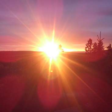 Das Bild zeigt einen atemberaubenden Sonnenaufgang über einer ruhigen Landschaft. Die Strahlen der Sonne erleuchten den Himmel und werfen ein warmes Licht auf die umliegenden Hügel und Bäume.