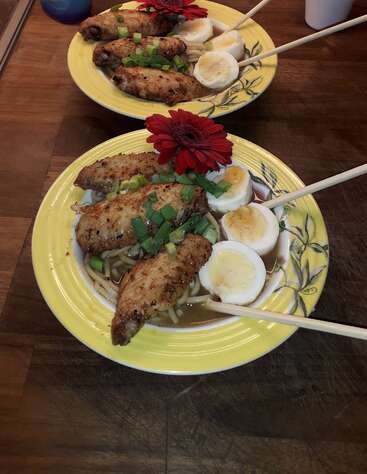 La imagen muestra dos cuencos de ramen sobre una mesa de madera, cada uno con fideos, alitas de pollo, huevos cocidos y adornados con cebollas verdes y una flor roja.