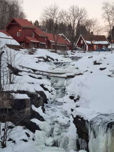 Un pintoresco pueblo de casas rojas se asienta junto a un arroyo parcialmente helado. La nieve cubre el suelo, los árboles están desnudos y el invierno domina el apacible paisaje.