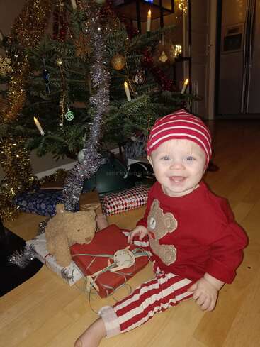 Ein fröhliches Baby in einem festlichen Schlafanzug sitzt neben einem geschmückten Weihnachtsbaum mit eingepackten Geschenken und einem Teddybär und lächelt strahlend in einem gemütlichen, warm beleuchteten Zimmer.