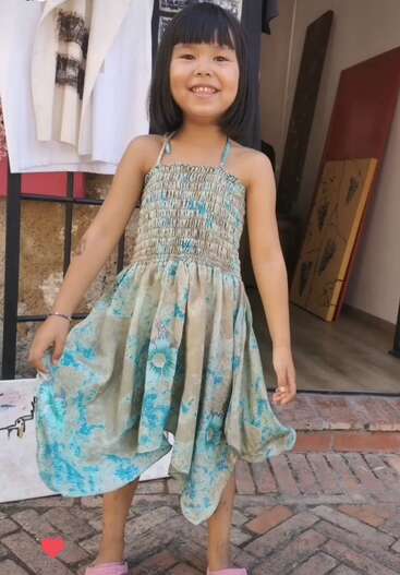 The image depicts a young girl with dark hair and bangs, wearing a light blue dress with a floral pattern, standing in front of a clothing rack and a wall.