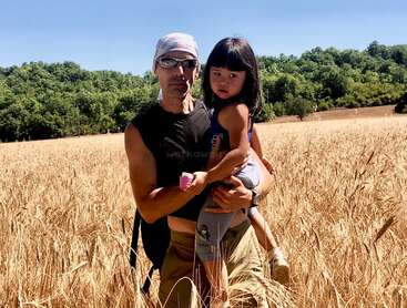 The image depicts a man holding a young girl in a wheat field, with trees visible in the background and a clear blue sky above.