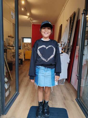 A smiling child stands in a shop entrance, wearing a black heart sweatshirt, denim skirt, black boots, and a cap. The shop displays art and clothing.