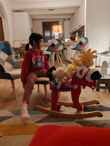 A young child sits on a chair in a cozy, warmly-lit living room. Stuffed animals are arranged on a red rocking horse in front.