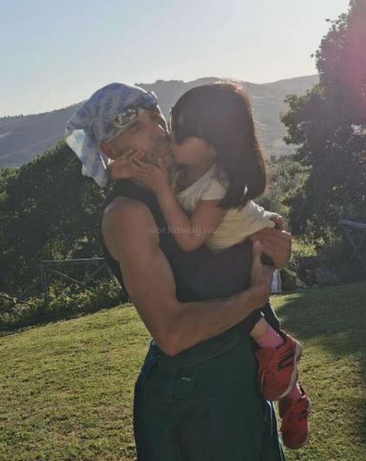 A man in a black tank top and headscarf is holding a child in his arms, kissing the child\'s cheek in a grassy field.