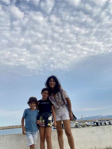 A woman with two children stands outdoors by the sea, smiling. The sky is filled with puffy clouds, and boats are visible in the background.