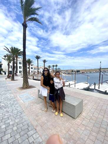 A woman and two children pose by a marina with boats, palm trees, and a blue sky with clouds. They appear happy, enjoying a scenic day.