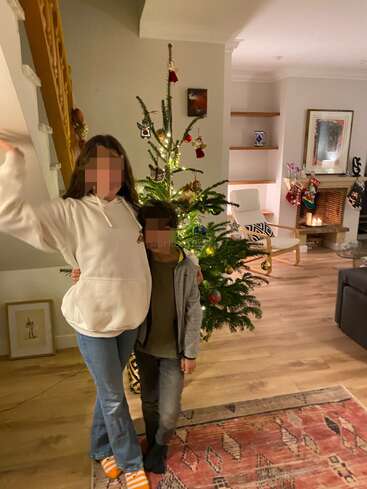 A woman and child pose near a decorated Christmas tree in a cozy living room, with stockings on the fireplace and warm festive decorations around.