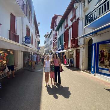 Una soleada escena callejera en una ciudad europea, con gente comprando y paseando. Edificios coloridos con contraventanas se alinean a lo largo de la calle. Tres personas posan felices en el centro.