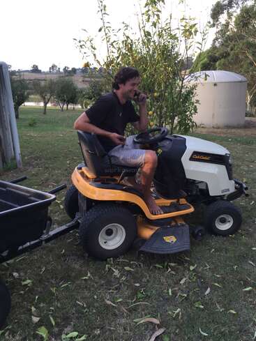 A man sits barefoot on a yellow lawn mower, talking on a phone, in a grassy yard with trees, a water tank, and scattered leaves.