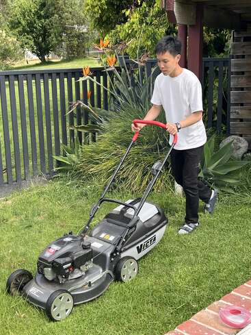 A young person wearing a white t-shirt and black pants is mowing the lawn with a Victa lawnmower in a green garden near a black fence.