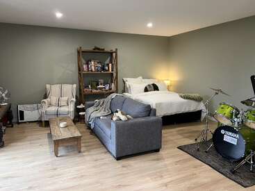 A cozy bedroom features a bed, armchair, small sofa, coffee table, bookshelf, lamp, and a green Yamaha drum set on a hardwood floor.