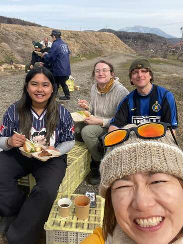 A group of friends sit outdoors, smiling and enjoying a meal together. Mountains and countryside are in the background. The atmosphere is cheerful, relaxed, and casual.