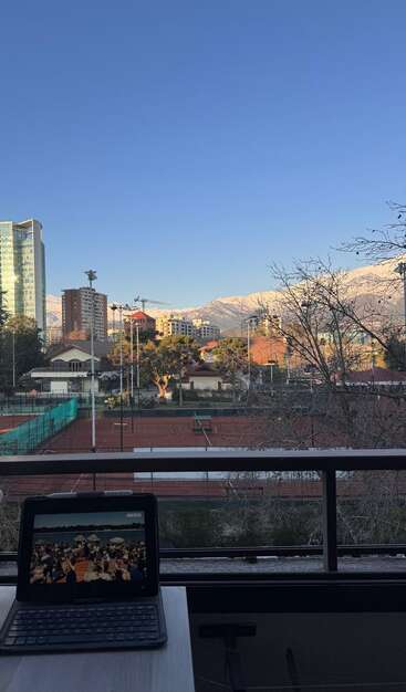 A tablet sits on a table facing red tennis courts, city buildings, and snowy mountains under a clear blue sky, blending technology with scenic tranquility.