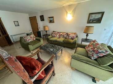 This cozy living room features green sofas with floral cushions, a marble coffee table, armchair, two lamps, paintings on the walls, and cream tiled floor.