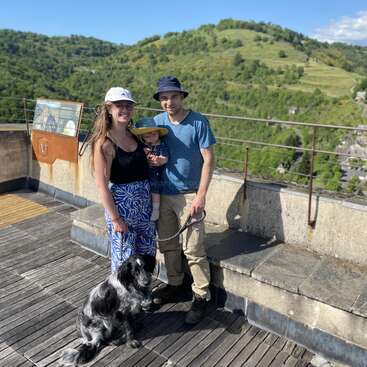 Une famille souriante accompagnée d'un chien noir et blanc se tient sur un point de vue ensoleillé, surplombant des collines verdoyantes et un ciel bleu clair, profitant d'une belle journée en plein air.