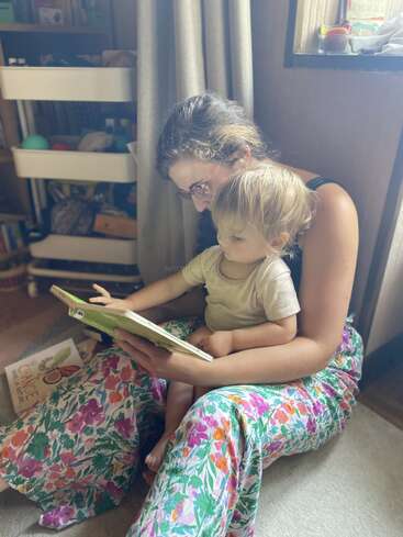 Una mujer sentada en el suelo con un niño en el regazo. Leen juntos un libro, rodeados de libros y de una luz suave y acogedora.