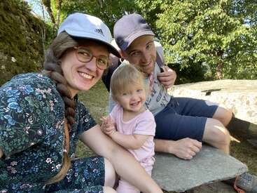 Una familia feliz está sentada al aire libre, sonriendo a la cámara. La mujer lleva gafas y gorra, el hombre se arrodilla junto a ellos y un alegre niño pequeño ríe.