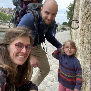A cheerful family of three, including a small child with glasses, poses happily by a riverside stone path, enjoying a day outdoors near a historic bridge.