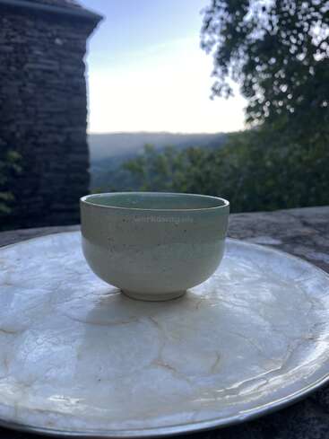 Uma xícara de cerâmica está sobre um prato grande e redondo ao ar livre. Ao fundo, um prédio de pedra, árvores e colinas distantes são suavemente iluminados pela luz do entardecer. Um clima tranquilo.