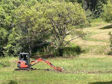 A red excavator works on a green grassy field, digging a trench. Surrounded by lush trees and sunlight, the peaceful landscape shows nature and machinery together.