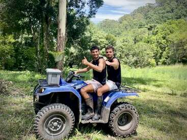 Two young men sit on a blue ATV in a lush, green outdoor setting. Both are smiling, giving thumbs up, wearing boots, sleeveless shirts, and shorts.