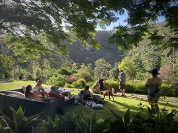 A group of people relax outdoors on comfortable furniture, surrounded by lush greenery, mountains, and sunshine. Some interact with a dog; the mood is peaceful and social.