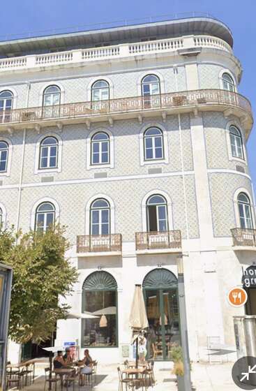 Ein schönes altes Gebäude mit gewölbten Fenstern, dekorativen Balkonen und einer Kachelfassade. Cafétische im Freien und Menschen, die einen sonnigen Tag unter Sonnenschirmen genießen, verleihen dem Gebäude Charme.